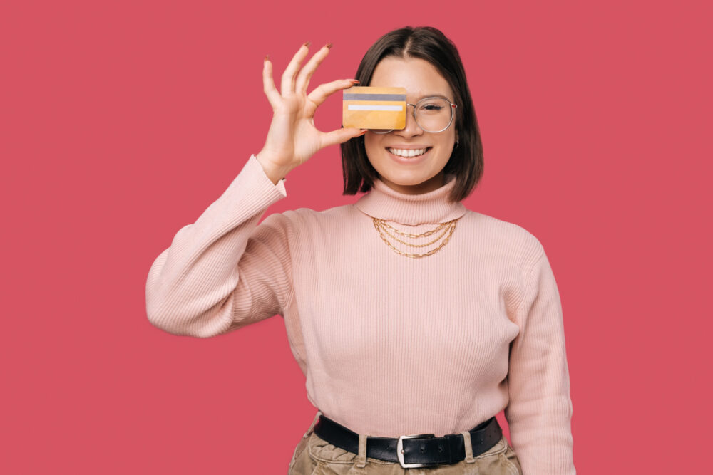 Photo of joyful young trendy woman holding yellow credit card over eyes and looking at the camera.