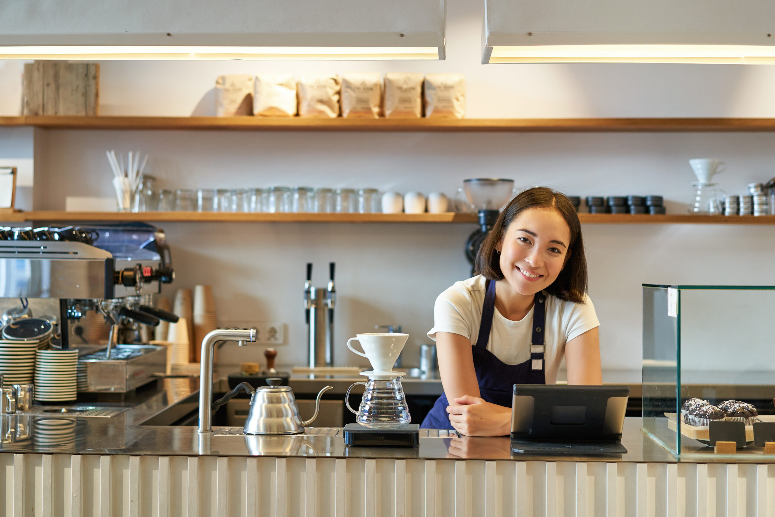 Happy smiling asian barista, girl behind counter, working with POS terminal and brewing filter kit, making coffee in cafe.